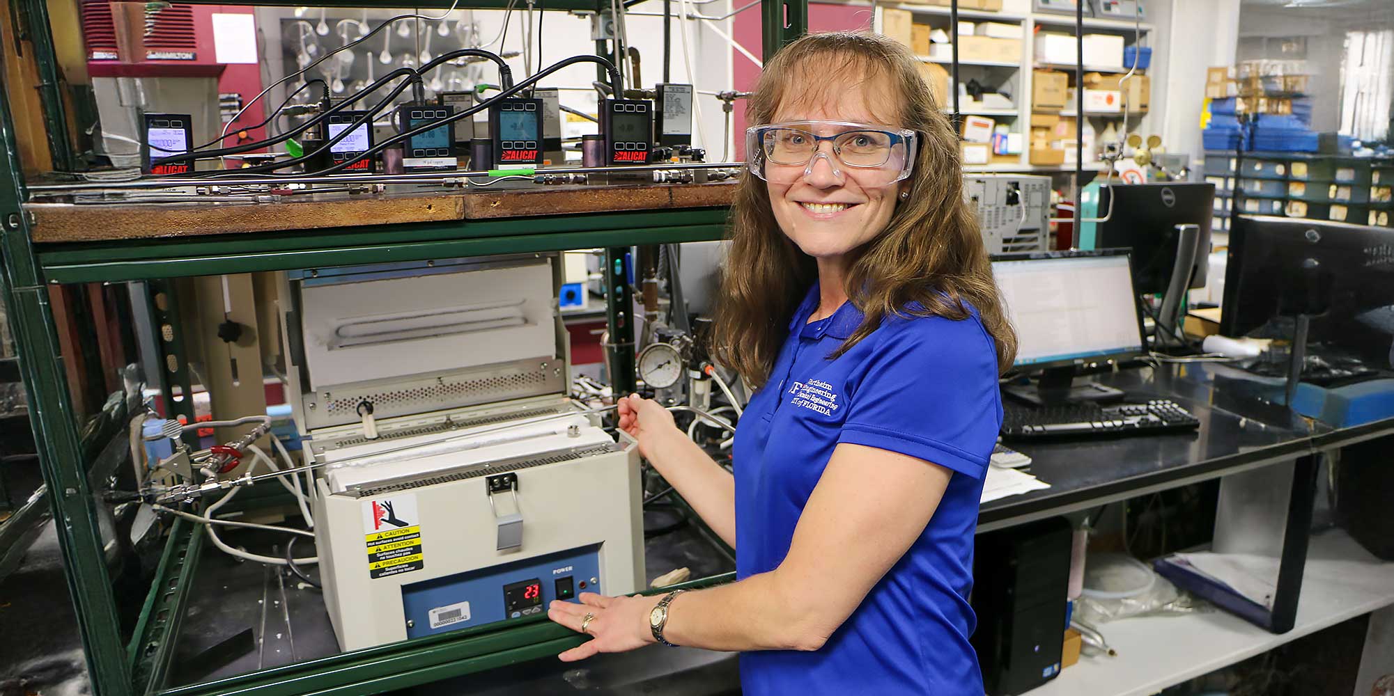 Helena Hagelin Weaver, Ph.D., working in a chemical engineering lab with catalyst testing equipment, wearing safety glasses and a blue UF polo.